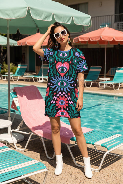 A short-sleeve, above-the-knee t-shirt dress with a true black background. The front features a vibrant, vertically stacked symmetrical graphic design. From top to bottom, the design includes a pink heart containing a green and blue eye, a large blue sunburst with a central eye, a red daisy flower with an eye flanked by pink and black checkered arches, and a pink mushroom cap with an eye. Swirling emerald green foliage, pink stars, and purple stars fill the negative space around the motifs.