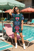 A short-sleeve, above-the-knee t-shirt dress with a true black background. The front features a vibrant, vertically stacked symmetrical graphic design. From top to bottom, the design includes a pink heart containing a green and blue eye, a large blue sunburst with a central eye, a red daisy flower with an eye flanked by pink and black checkered arches, and a pink mushroom cap with an eye. Swirling emerald green foliage, pink stars, and purple stars fill the negative space around the motifs.