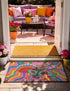 A rectangular doormat featuring a vibrant 2D flat print designed to look like textured yarn embroidery. The central graphic showcases a bright green alien with large black eyes, wearing a pink neck bandana and a highly decorated red, pink, and blue sombrero cowboy hat. The alien has one green hand raised in a friendly wave. The background is a psychedelic pattern of wavy, flowing rainbow stripes in yellow, orange, red, pink, blue, and green. The mat has a sleek black outer edge trim.