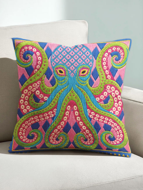 A square decorative throw pillow cover featuring a highly symmetrical bright octopus design. The octopus has a turquoise blue body with a diamond patterned head and eight curled tentacles detailed in bright pink and lime green. The background is a repeating diamond argyle pattern in royal blue and light pink. The artwork is a flat two dimensional print that mimics the textured look of intricate crewelwork embroidery. The pillow features a hidden zipper on the bottom edge. No insert included.