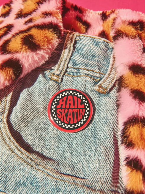 A round red pinback button with a glossy, scratch-resistant mylar face. The design features the phrase "HAIL SKATIN'" in a wavy, 70s psychedelic font in the center. This text is surrounded by a bold, black and white checkerboard pattern border, giving it a punk rock aesthetic. This piece of vintage-inspired flair is made of lightweight metal with a durable safety pin backing on the reverse. The button is a perfect for a denim jacket, backpack, or bag for fans of roller derby and skateboarding.