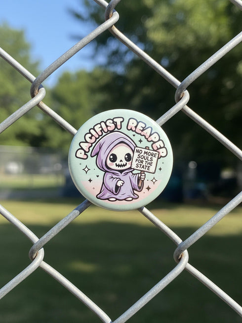 Round pinback button featuring a pastel mint green background with a cartoon grim reaper character in a lavender hood holding a protest sign. The design includes bold black outlined lettering reading “Pacifist Reaper” above the figure and small sparkle accents surrounding it. The finish is glossy and smooth, with a printed illustration style on a metal pin badge surface.