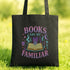 A black tote bag with a colorful graphic design on it, featuring the text "BOOKS ARE MY FAMILIAR" and various symbols related to books and reading.