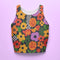 Vibrant retro cropped tank top featuring psychedelic 70s floral pattern with orange yellow purple flowers, black and white checkerboard accents, and wavy green lines on dark background. Body-hugging racerback design made from performance stretch fabric with moisture-wicking properties and UV protection. Perfect for festivals, raves, summer concerts, or bold streetwear styling.