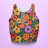 Vibrant retro cropped tank top featuring psychedelic 70s floral pattern with orange yellow purple flowers, black and white checkerboard accents, and wavy green lines on dark background. Body-hugging racerback design made from performance stretch fabric with moisture-wicking properties and UV protection. Perfect for festivals, raves, summer concerts, or bold streetwear styling.
