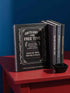 An 8.5 x 11 inch black hardcover spiral planner with a durable satin matte laminated finish. The front cover features a vintage-style white ornamental border, creating a gothic, macabre aesthetic. Inside the frame, the title "Calendar of Doom" is printed in an antique serif font. This undated weekly and monthly agenda is shown with a metal wire-o spiral binding on the right side. The design uses a black, white, and red color scheme, making it a unique piece of dark humor stationery