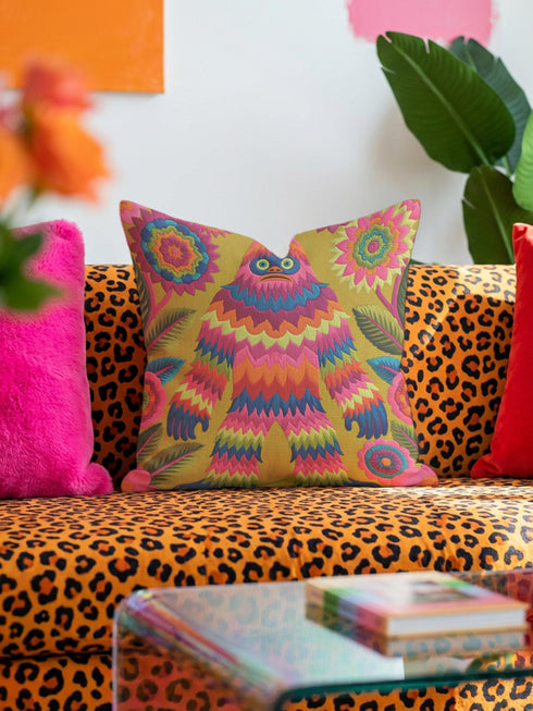 A square faux embroidery pillow featuring a brightly colored maximalist design against a mustard yellow background. In the center stands a stylized, furry Bigfoot or Sasquatch figure rendered in vibrant layers of pink, orange, blue, and lime green with a blue face and yellow eyes. The central creature is surrounded by large, detailed symmetrical pink and blue flowers and leaves. The flat printed fabric creates a highly realistic optical illusion of dimensional crewel embroidery stitches.