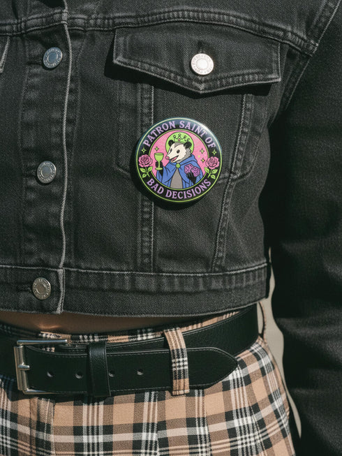 A round glossy metal pinback button featuring a flat 2D print illustration of an opossum wearing a blue cloak and a green crown. The opossum holds a neon green goblet in one paw and two purple dice in the other. A bright pink circle sits behind the opossum, surrounded by a black outer ring. Purple text reads PATRON SAINT OF BAD DECISIONS curved around the edge. Two pink roses with green leaves are on the sides. The design uses bold black outlines and bright neon colors.