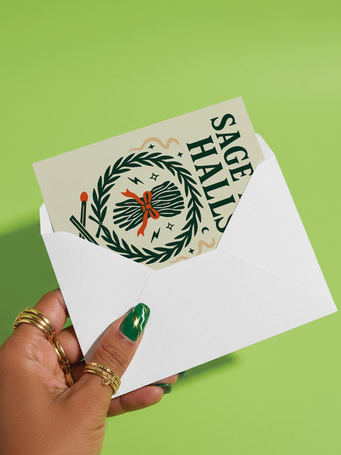 A detailed folk-art style illustration of a "Sage the Halls" holiday greeting card. The design features a bundle of sage smudge sticks tied with a red ribbon at the center of a dark green wreath. The wreath is accented with small crescent moons, stars, and smoke swirls. Below the wreath are two crossed matches with red tips. The card is printed on creamy beige paper with bold, dark green serif text. This funny spiritual and witchy Yule card is a unique pagan greeting for energy cleansing.