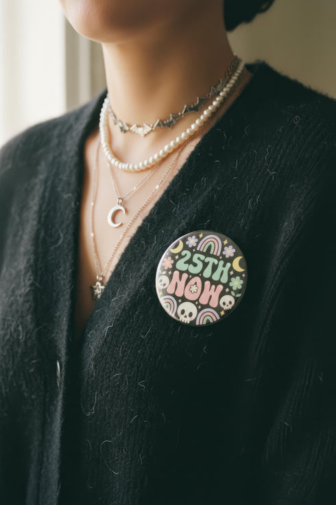 A round pinback button featuring a solid black background covered in a retro pastel colored design. The center reads 25TH NOW in a groovy bubbly font using pale pink and mint green referencing the 25th amendment. Surrounding the text is a maximalist cluster of cute but edgy motifs including smiling white skulls, pastel rainbows, pale yellow crescent moons, daisies, and a pink and white spotted mushroom. The button has a glossy laminate finish and a secure metal safety pin backing.