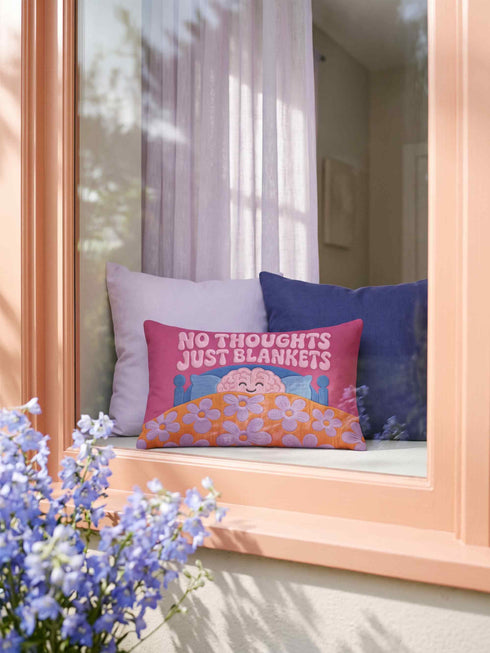 A bright magenta pink rectangular lumbar pillow cover featuring a flat 2D printed faux embroidery design. The printed illusion shows light pink bubble text reading NO THOUGHTS JUST BLANKETS. Below the text is a cartoon pink brain with a happy face, tucked into a blue bed with blue pillows. The brain is covered by a textured-looking orange blanket decorated with light purple daisies. The entire design is a smooth printed trompe l'oeil stitch effect, not real woven or stitched texture.