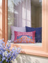 A bright magenta pink rectangular lumbar pillow cover featuring a flat 2D printed faux embroidery design. The printed illusion shows light pink bubble text reading NO THOUGHTS JUST BLANKETS. Below the text is a cartoon pink brain with a happy face, tucked into a blue bed with blue pillows. The brain is covered by a textured-looking orange blanket decorated with light purple daisies. The entire design is a smooth printed trompe l'oeil stitch effect, not real woven or stitched texture.
