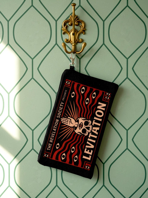 A rectangular makeup bag featuring a vibrant psychedelic print with gothic elements. The fabric texture is clearly visible showing a woven linen-like polyester grain. A black zipper runs across the top edge with a black detachable nylon wrist strap clip on the side, showcasing the approximately nine by six inch dimensions. The print includes high contrast neon colors and trippy swirling patterns typical of maximalist decor.