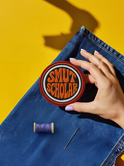 A hand holding a circular patch with the text "SMUT SCHOLAR" embroidered on it, along with a spool of thread and a needle, all placed on a blue denim fabric against a yellow background.