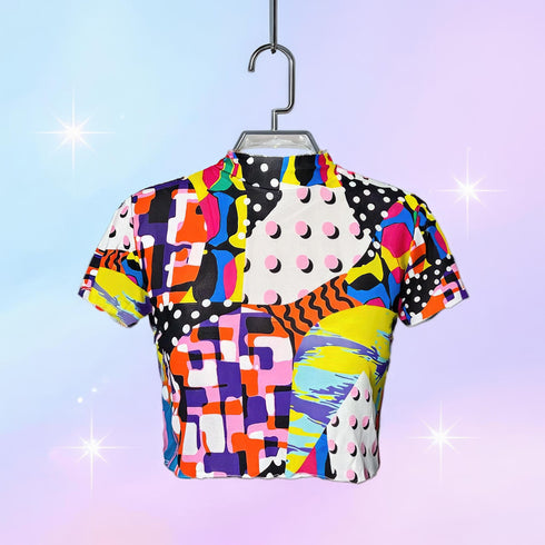 A colorful 90s-style mesh crop top featuring a maximalist abstract pattern. This see-through shirt has a high mock neck, short sleeves, and a vibrant, retro design with geometric shapes, color blocking, and polka dots in bold shades of pink, yellow, blue, and orange. The Y2K-inspired top is a statement piece of festival clothing, perfect for a rave outfit or alternative, dopamine-dressing-inspired streetwear looks.