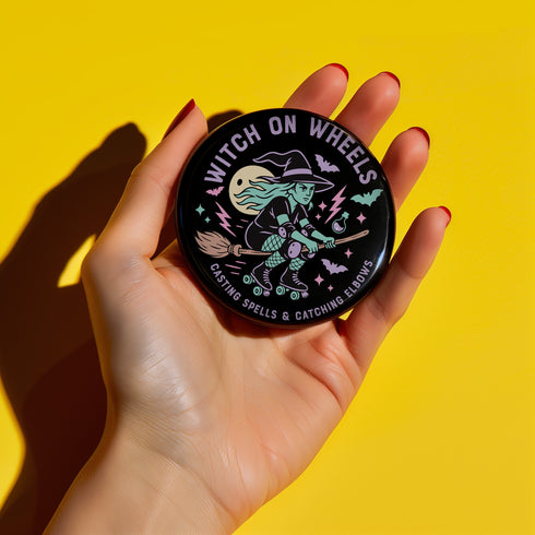 A hand holding a round pin with a witch riding a broomstick and the text "Witch on Wheels" printed on it.