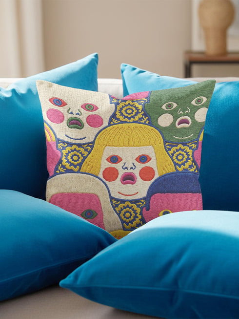 A square decorative pillow cover featuring a faux embroidery pattern of five stylized, expressive doll faces. The central face has yellow hair and pink cheeks. It is surrounded by four other faces in shades of pale beige, green, and bright pink, all with wide staring eyes and open mouths. The design uses intricate, colorful line work to create the illusion of thick woven stitching, but the fabric is a smooth print. Medallions fill the gaps.