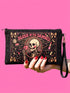 A rectangular gothic makeup bag made of black polyester with a visible woven linen-like crosshatch texture. The central illustration features a smiling human skull with flowing hair applying a pink heart-shaped blush to its cheek. Above the skull, the text BLUSH OF THE DAMNED appears. The design is framed by illustrations of lipstick tubes, mascara wands, bows. The bag includes a black zipper along the top edge and a removable black nylon wrist strap attached to the metal zipper pull.