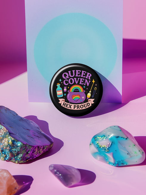 Glossy black pinback button with pastel artwork of a bubbling cauldron, rainbow, potion bottles, and tools. Text reads “Queer Coven, Hex Proud.”