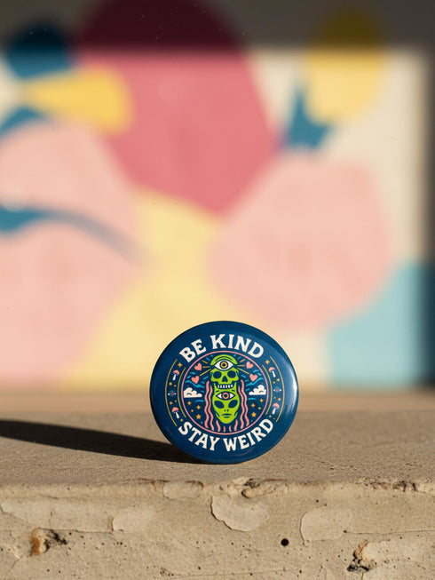 A round 1.75 inch pinback button featuring a retro psychedelic design on a dark navy blue background. The text reads BE KIND STAY WEIRD in vintage cream lettering around the border. The center illustration depicts a neon green skull stacked on top of a neon green alien head. Both the skull and alien have opening third eyes on their foreheads. Pink wavy lines frame the faces. The background is decorated with small colorful mushrooms, hearts, stars, and clouds. Glossy finish.