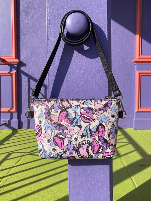 Small vegan leather crossbody purse with a real pebble grain texture and adjustable black strap. The flat printed artwork features a scattered all over pattern of pink skulls, purple and pastel blue winged bats, pink fanged mouths, and loose eyeballs set against a cream colored background. The bag is shown with black and silver hardware including a black top zipper, black zipper pull, black strap tabs, and silver metal rings and clips. The compact rectangular bag measures 8.5 by 7 by 2 inches.