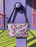Small vegan leather crossbody purse with a real pebble grain texture and adjustable black strap. The flat printed artwork features a scattered all over pattern of pink skulls, purple and pastel blue winged bats, pink fanged mouths, and loose eyeballs set against a cream colored background. The bag is shown with black and silver hardware including a black top zipper, black zipper pull, black strap tabs, and silver metal rings and clips. The compact rectangular bag measures 8.5 by 7 by 2 inches.