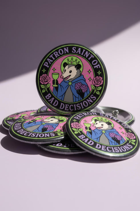 A round glossy metal pinback button featuring a flat 2D print illustration of an opossum wearing a blue cloak and a green crown. The opossum holds a neon green goblet in one paw and two purple dice in the other. A bright pink circle sits behind the opossum, surrounded by a black outer ring. Purple text reads PATRON SAINT OF BAD DECISIONS curved around the edge. Two pink roses with green leaves are on the sides. The design uses bold black outlines and bright neon colors.