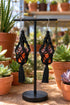 A pair of black crochet earrings with orange pots containing succulent plants, displayed on a black stand in a greenhouse-like setting with various potted plants in the background.