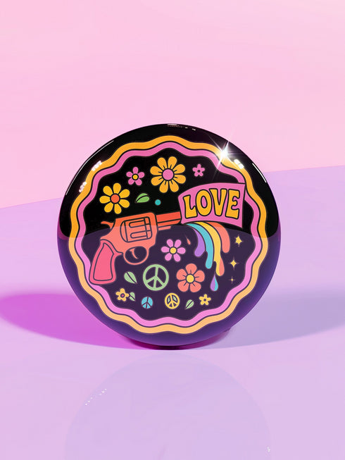 Round pinback button badge with a black background, featuring a retro illustration of a red revolver shooting out a rainbow flag that says “LOVE.” Surrounded by colorful 70s-style flowers and peace signs, outlined in wavy neon border. Glossy finish with vibrant pop colors and psychedelic design.