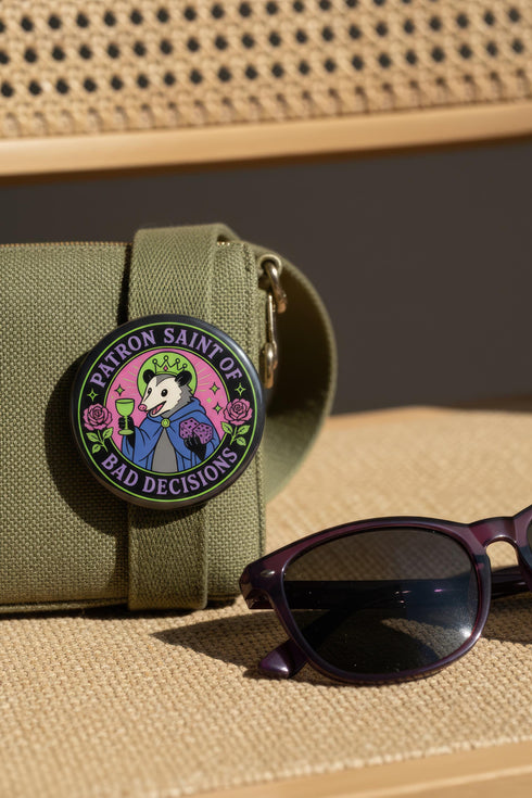 A round glossy metal pinback button featuring a flat 2D print illustration of an opossum wearing a blue cloak and a green crown. The opossum holds a neon green goblet in one paw and two purple dice in the other. A bright pink circle sits behind the opossum, surrounded by a black outer ring. Purple text reads PATRON SAINT OF BAD DECISIONS curved around the edge. Two pink roses with green leaves are on the sides. The design uses bold black outlines and bright neon colors.
