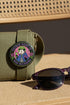 A round glossy metal pinback button featuring a flat 2D print illustration of an opossum wearing a blue cloak and a green crown. The opossum holds a neon green goblet in one paw and two purple dice in the other. A bright pink circle sits behind the opossum, surrounded by a black outer ring. Purple text reads PATRON SAINT OF BAD DECISIONS curved around the edge. Two pink roses with green leaves are on the sides. The design uses bold black outlines and bright neon colors.