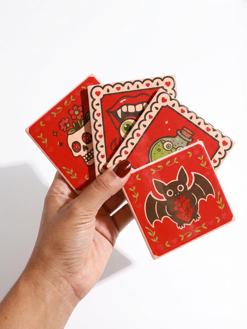 Set of four square glazed ceramic coasters featuring a vibrant pastel goth and creepy cute design. These functional art pieces show a spooky yet playful aesthetic perfect for alternative home decor. The coasters have a glossy wipe-clean finish and a non-slip cork backing to protect furniture surfaces from hot and cold beverages. Ideal for maximalist coffee tables, witchy desks, or as a unique housewarming gift for horror fans who love retro 70s and dopamine decor styles.