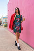 A short-sleeve, above-the-knee t-shirt dress with a true black background. The front features a vibrant, vertically stacked symmetrical graphic design. From top to bottom, the design includes a pink heart containing a green and blue eye, a large blue sunburst with a central eye, a red daisy flower with an eye flanked by pink and black checkered arches, and a pink mushroom cap with an eye. Swirling emerald green foliage, pink stars, and purple stars fill the negative space around the motifs.