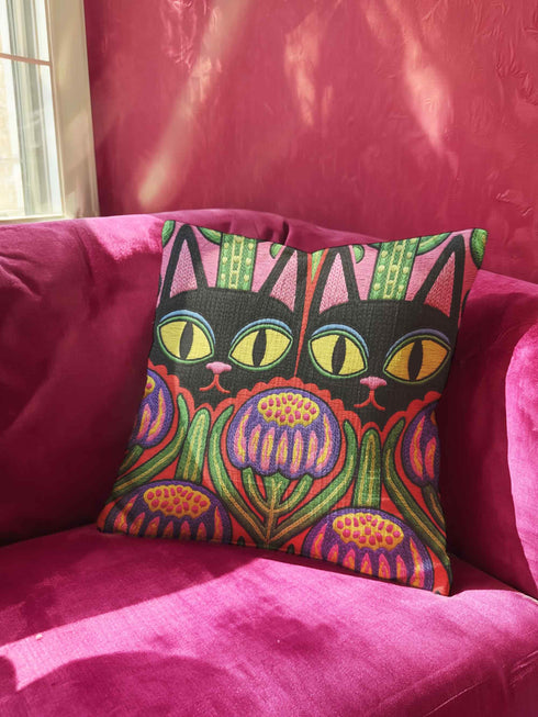 A square decorative pillow cover featuring a vibrant, symmetrical flat printed design that mimics the look of textured embroidery. Two black cats with large yellow eyes and pink noses face forward against a bright red background. Below the cats are large purple and pink flowers with green stems. Above the cats are green cactus shapes. The artwork uses bold, colorful outlines to create an optical illusion of stitching, though the fabric itself is smooth.