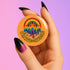 Round orange pinback button featuring bold rainbow psychedelic artwork of a melting third-eye face beneath a rainbow arch. Surrounded by stars and the phrase “Hot, Queer and Radiating” in retro black bubble letters. Glossy metal badge with UV-resistant mylar face and secure safety pin back.