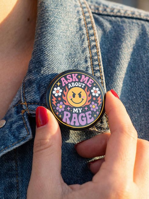 A round glossy metal pinback button featuring a flat 2D print design on a solid black background. In the center is an angry yellow smiley face surrounded by orange and red flames. Above the smiley face, pink text reads ASK ME and white text reads ABOUT. Below the face, white text reads MY and large purple text reads RAGE. The design is accented with two white daisies, two purple flowers, pink and orange four-point stars, and orange lightning bolts framing the outer edges.