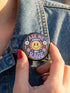 A round glossy metal pinback button featuring a flat 2D print design on a solid black background. In the center is an angry yellow smiley face surrounded by orange and red flames. Above the smiley face, pink text reads ASK ME and white text reads ABOUT. Below the face, white text reads MY and large purple text reads RAGE. The design is accented with two white daisies, two purple flowers, pink and orange four-point stars, and orange lightning bolts framing the outer edges.