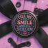 Glossy black pinback button with pastel-colored embroidered-style text that reads “Tell Me to Smile and I’ll Scream,” with lips icon and star accents.