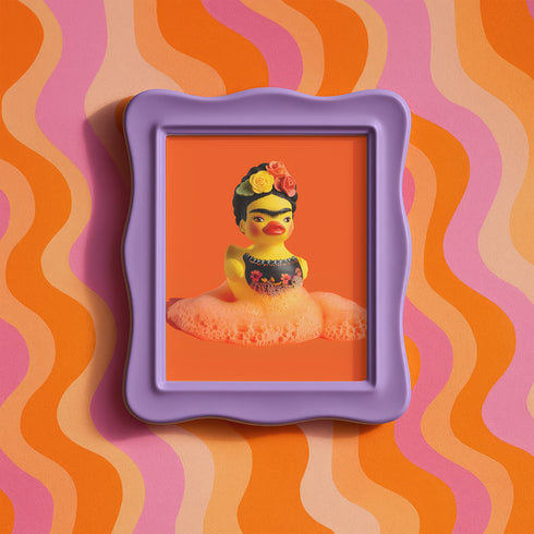 A vibrant fine art print of a rubber duck inspired by Frida Kahlo, complete with bold unibrow, floral crown, and traditional embroidered blouse. Framed in a red sculptural frame, the duck floats in orange-tinted bubbles against a deep orange backdrop and violet wall.
