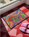A rectangular doormat featuring a vibrant 2D flat print designed to look like textured yarn embroidery. The central graphic showcases a bright green alien with large black eyes, wearing a pink neck bandana and a highly decorated red, pink, and blue sombrero cowboy hat. The alien has one green hand raised in a friendly wave. The background is a psychedelic pattern of wavy, flowing rainbow stripes in yellow, orange, red, pink, blue, and green. The mat has a sleek black outer edge trim.
