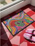 A rectangular doormat featuring a vibrant 2D flat print designed to look like textured yarn embroidery. The central graphic showcases a bright green alien with large black eyes, wearing a pink neck bandana and a highly decorated red, pink, and blue sombrero cowboy hat. The alien has one green hand raised in a friendly wave. The background is a psychedelic pattern of wavy, flowing rainbow stripes in yellow, orange, red, pink, blue, and green. The mat has a sleek black outer edge trim.