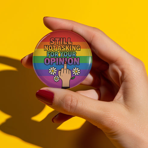 Round glossy pinback button measuring 2.25 inches with a bold rainbow pride flag background. Centered text reads “Still Not Asking For Your Opinion” in black, yellow, blue, and purple letters. Below the text is an illustrated hand with painted red nails giving the middle finger, surrounded by white daisies. The design is printed on tinplate with a scratch- and UV-resistant mylar coating. Button features a strong safety pin backing, perfect for bags, jackets, or pride outfits.