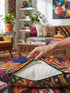 A square decorative throw pillow cover featuring a trippy retro Bigfoot scene. A large navy blue silhouette of Sasquatch walks through a forest of navy and yellow pine trees. The background is a psychedelic swirling sky in vibrant pink and yellow, featuring a large stylized eye and a cluster of small yellow stars. The flat two dimensional print visually mimics the textured look of heavy needlepoint embroidery. The pillow features a sleek hidden zipper on the bottom. No insert is included.