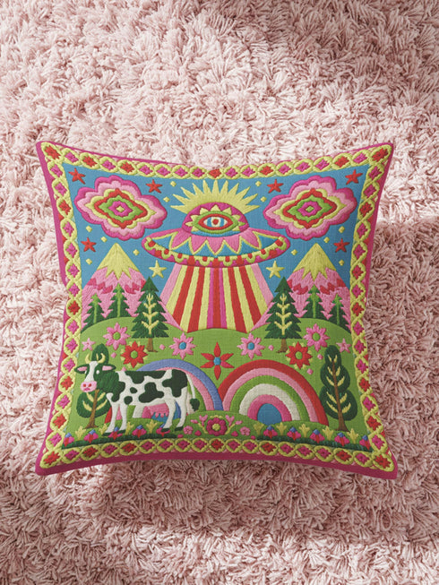 Description of a square throw pillow with a vibrant maximalist alien design. A central UFO with a large all-seeing eye hovers above a landscape with two rainbows, pink mountains, and green pine trees. A black and white spotted cow stands in the foreground. The design is a high-contrast palette of sky blue, magenta, hot pink, and meadow green. The pillow has a faux-embroidery 2D print finish on linen-feel fabric, framed by a yellow and pink scalloped floral border.