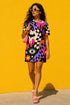A short-sleeve, above-the-knee t-shirt dress with a relaxed, straight fit and a crew neckline. The dress features a vibrant, chaotic, all-over graphic print. The design includes sections of distorted, wavy black and cream checkerboard, mixed with bold, abstract shapes in bright purple, hot pink, and vivid orange. Scattered throughout the pattern are motifs of hot pink dripping slime, a black bat silhouette, a stylized green eye with a pink iris, and retro pink and purple daisy flowers.