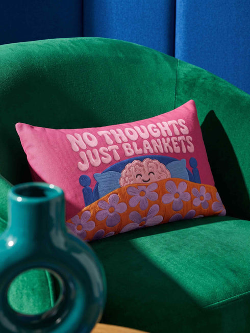 A bright magenta pink rectangular lumbar pillow cover featuring a flat 2D printed faux embroidery design. The printed illusion shows light pink bubble text reading NO THOUGHTS JUST BLANKETS. Below the text is a cartoon pink brain with a happy face, tucked into a blue bed with blue pillows. The brain is covered by a textured-looking orange blanket decorated with light purple daisies. The entire design is a smooth printed trompe l'oeil stitch effect, not real woven or stitched texture.