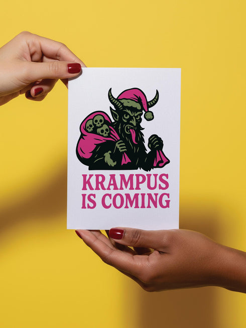 A 5x7 inch Krampus Christmas card shown in two paper finishes. The front features a stylized illustration of the folklore demon Krampus with green skin, black horns, and a pink Santa hat. He carries a large pink sack filled with three skulls over his shoulder. Below him, the words "KRAMPUS IS COMING" are printed in bold pink text. The inside of the card is blank on the left and has the printed text "Hope you've been beautifully bad this year." on the right.
