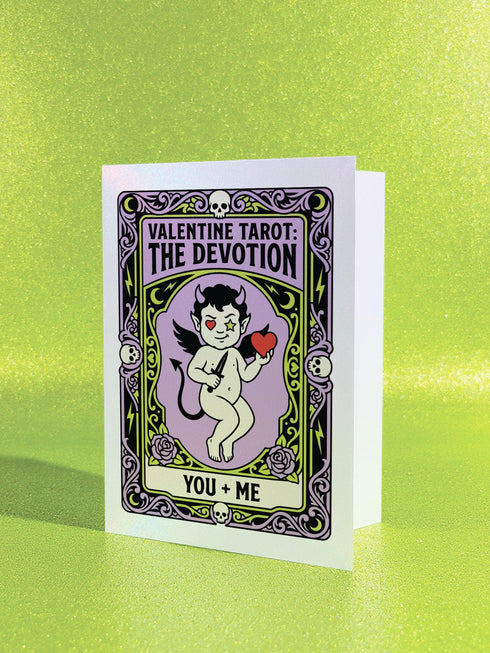 A vertical folded greeting card featuring a pastel goth tarot design titled The Devotion. The central illustration shows a pale devil imp with black wings and horns, holding a red heart in the left hand and a black dagger in the right. The imp has one star eye and one heart eye. The background is lavender purple. A black border frames the scene with teal vines, skulls, gold crescent moons, and roses. The bottom banner reads YOU + ME. The finish is shimmering pearlescent.