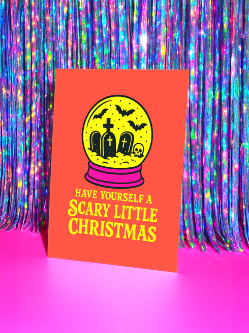 A red greeting card with a yellow snow globe design and the text "HAVE YOURSELF A SCARY LITTLE CHRISTMAS" on it, placed against a background of colorful, glittery curtains.
