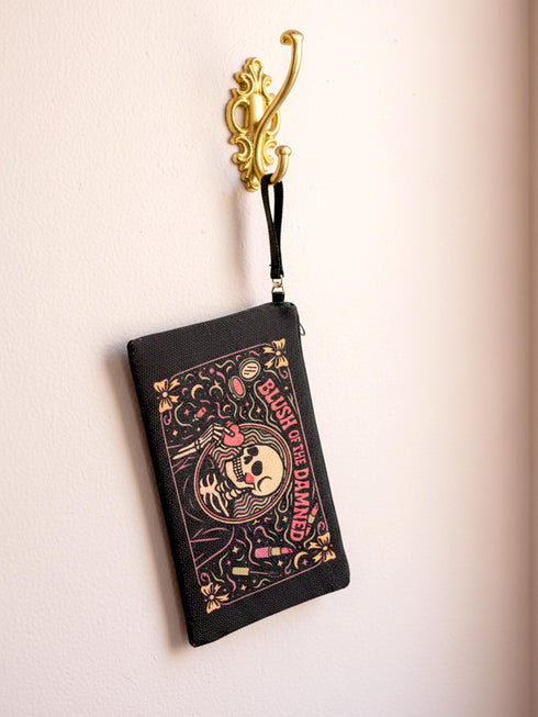 A rectangular gothic makeup bag made of black polyester with a visible woven linen-like crosshatch texture. The central illustration features a smiling human skull with flowing hair applying a pink heart-shaped blush to its cheek. Above the skull, the text BLUSH OF THE DAMNED appears. The design is framed by illustrations of lipstick tubes, mascara wands, bows. The bag includes a black zipper along the top edge and a removable black nylon wrist strap attached to the metal zipper pull.